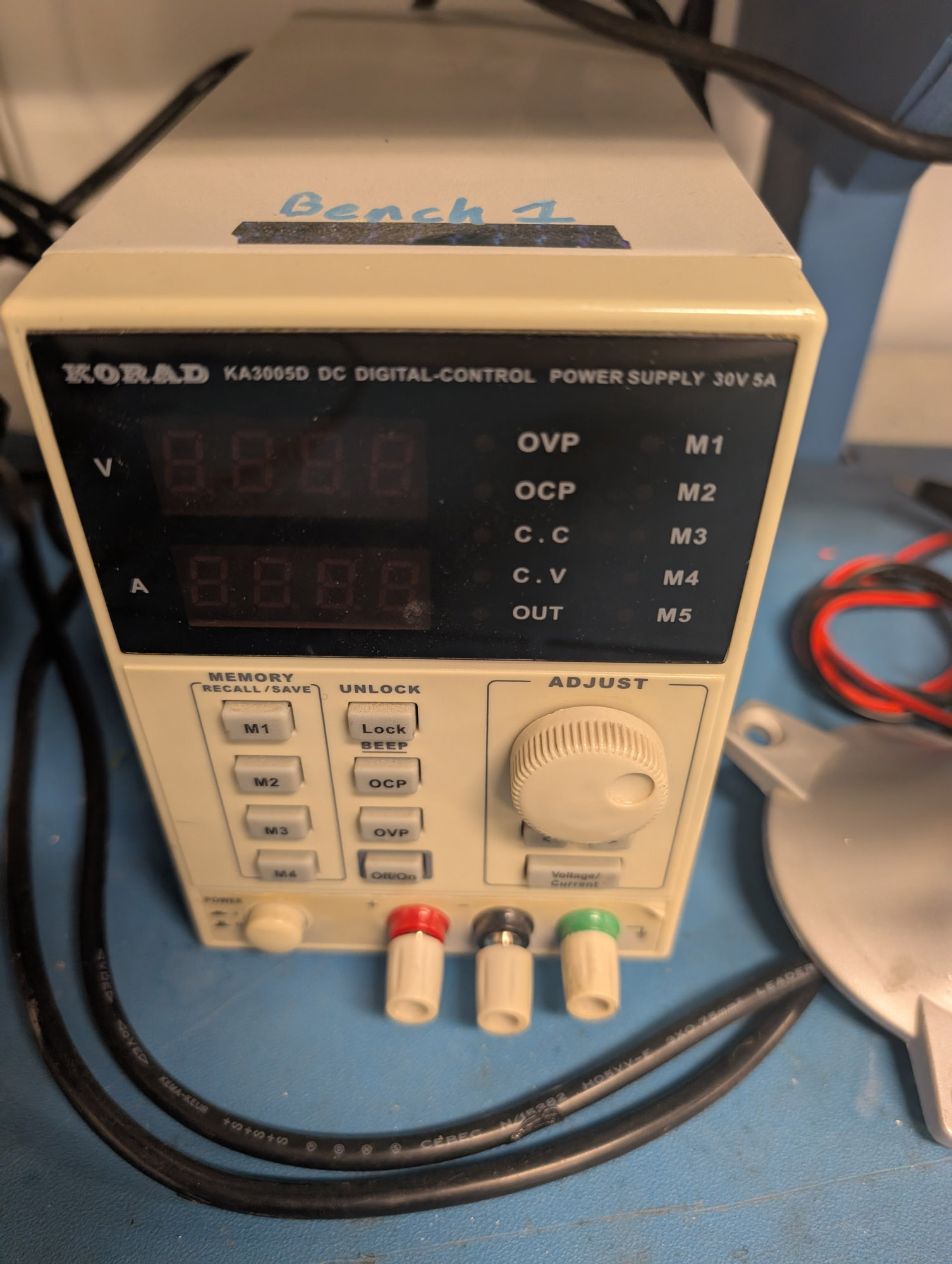A beige power supply, with “KORAD KA3005D DC Digital-Control Power Supply 30V 5A” across the top, 4-digit displays for V and A on the left, LEDs for OVP, OCP, CC, CV, Out, M1-M5 to the right of that. Four memory buttons as well as On/Off, Lock, OCP, and OVP buttons are below those, as well as a wheel labelled Adjust, and red, black, and green combo banana jack/binding posts at the bottom, along with a hard power button.